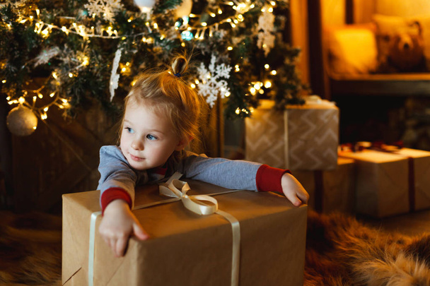 Een mooi klein meisje hugs een doos met een gift van Kerstmis voor een versierde kerstboom. Familie vriendelijke kerst concept. - Foto, afbeelding
