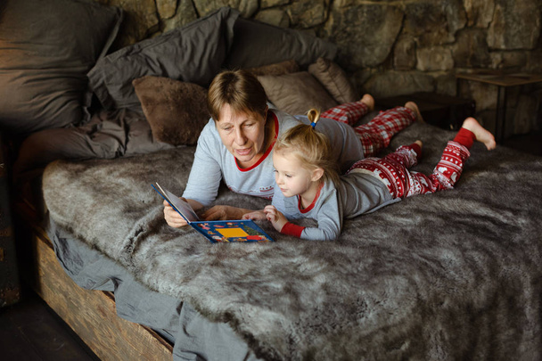 Бабушка и внучка веселятся вместе, читая книгу на кровати. Концепция семейного Рождества
. - Фото, изображение