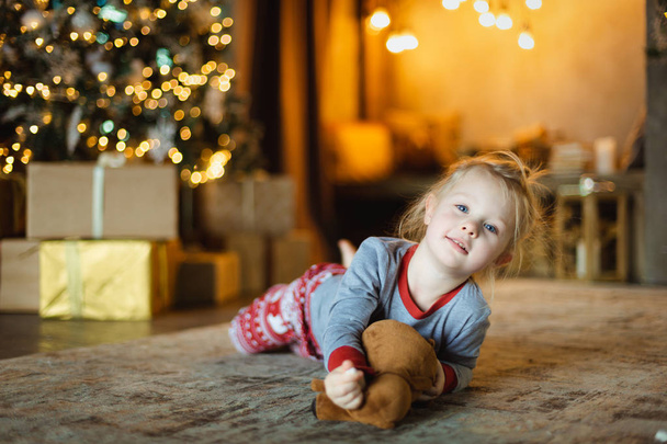 A beautiful baby lies on the floor in front of the tree and hugs her favorite plush toy - Valokuva, kuva