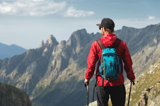 A bearded man in sunglasses and a cap with a backpack stands on top of a rock and looks into a rocky valley high in the mountains. The concept of tourism and easy trekking in the mountains outdoor - Fotoğraf, Görsel