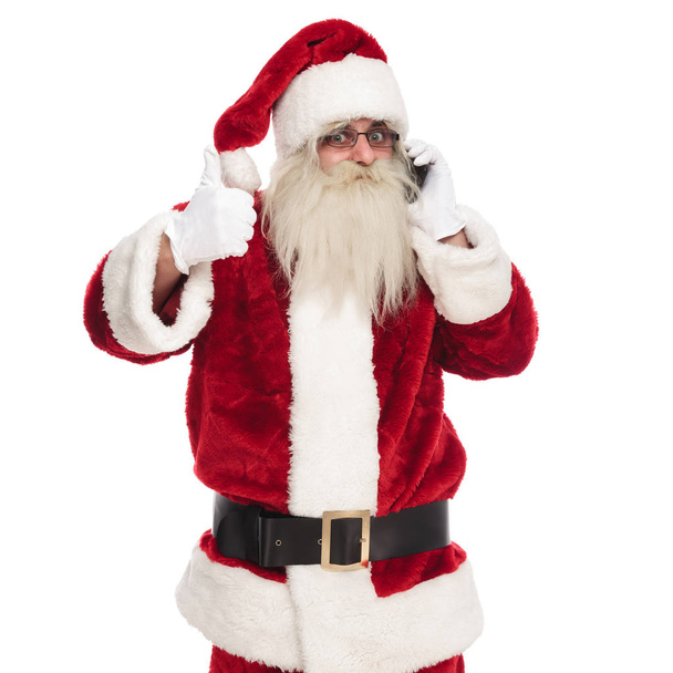 Porträt eines fröhlichen Weihnachtsmannes, der auf weißem Hintergrund telefoniert und ein Ok-Zeichen macht - Foto, Bild