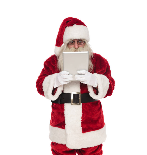 portrait of shocked santa looking at grey tablet while standing on white background - 写真・画像