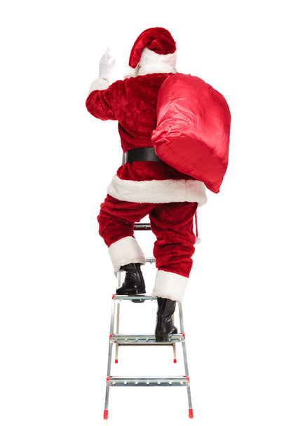 Rückansicht des Weihnachtsmannes mit Sack auf der Schulter, der auf einer Leiter auf weißem Hintergrund klettert, Bild in voller Länge - Foto, Bild