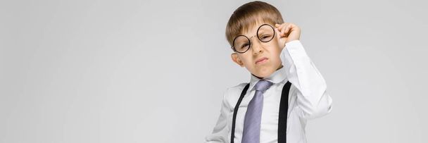 Un niño encantador con una camisa blanca, tirantes, corbata y pantalones vaqueros ligeros se levanta sobre un fondo gris. el niño levantó sus gafas
 - Foto, imagen