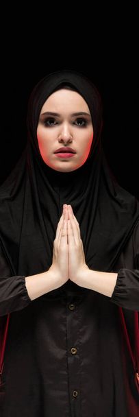 Portret van mooie ernstige jonge moslimvrouw dragen zwarte hijab met handen in de buurt van haar gezicht als bidden concept op zwarte achtergrond - Foto, afbeelding