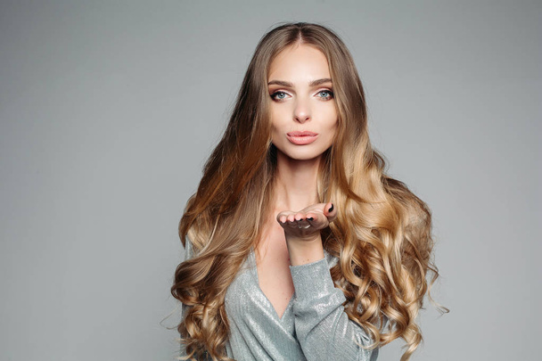 Studio portrait of an attractive blond woman with long thick hair and professional make-up wearing an elegant silver blouse, an air kiss on the camera on a gray background. Isolate. - Photo, Image