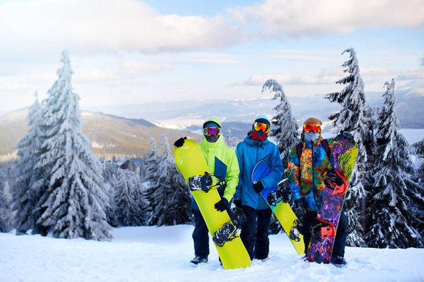 Tres snowboarders posando en la estación de esquí. Amigos subiendo a la cima de la montaña llevando sus tablas de snowboard a través del bosque para freeride backcountry y usando gafas reflectantes, ropa de moda colorida
. - Foto, imagen