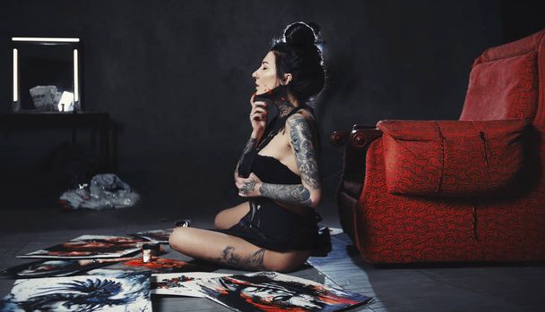 Maalattu taiteilija, jolla on tatuointi, istuu lattialla maalausten ja nojatuolin vieressä.
 - Valokuva, kuva
