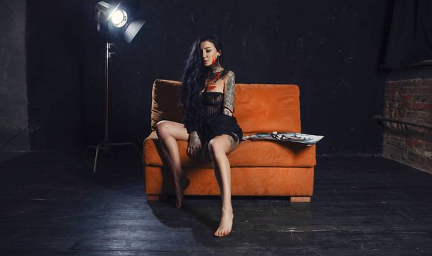 Dziewczyna z długimi włosami, siedząc na kanapie pomarańczowy. Dziewczyna, tatuaże ciała i farby - Zdjęcie, obraz
