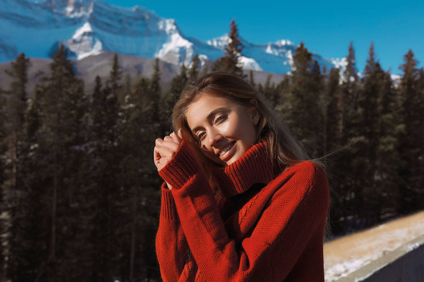 Inschrijving lachende model in zachte warme trui geniet haar wintervakantie in Alberta. De stralen van de zon vallen op haar gezicht, blonde haren schijnt. Natuur landschap rond, panoramisch uitzicht op de hoge bergen in de sneeuw - Foto, afbeelding