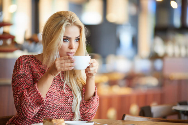 Blonde woman  looking through window while drinking tea sitting in cafe - Фото, изображение