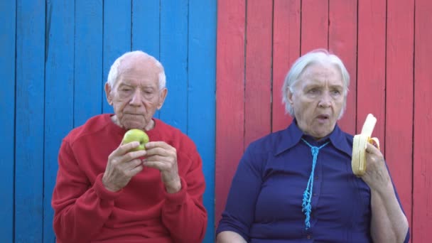 Συνταξιούχοι σε οικολογικά ξύλινα φόντο. Ο παππούς τρώει πράσινο μήλο και γιαγιά τρώει μπανάνα. Οι κινήσεις τους είναι αργή. Μοναδικό οικολογικό ξύλινο υπόβαθρο των δύο χρώματα (ροζ και μπλε). - Πλάνα, βίντεο