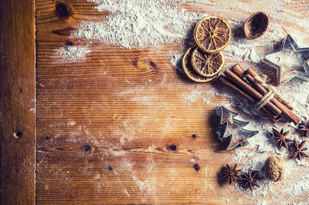 Top of view mesa de panadería llena de ingredientes aromáticos de Navidad anís estrella canela harina de naranja seca libro de cocina y cortadores de galletas
.  - Foto, imagen