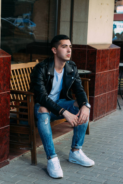 attractive guy sitting on a bench in a cafe on the street, white sneakers, white T-shirt, black leather jacket and jeans, pants. model. waiting for a meeting. - Fotó, kép
