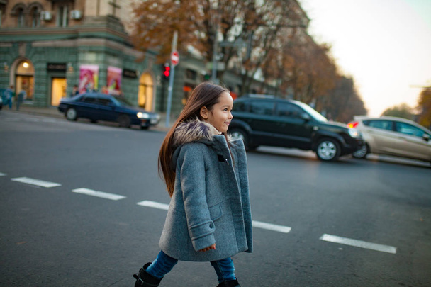 Dziecko dziewczynka w płaszcz jest chodzenie po drugiej stronie ulicy, na przejściu dla pieszych na tle miasta ulicy w godzinach wieczornych. - Zdjęcie, obraz