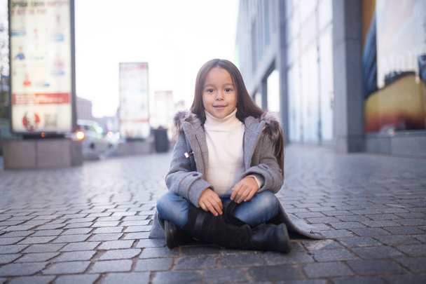 Child girl in a coat is sitting and smiling in the middle of the sidewalk on city street in evening. - Фото, изображение