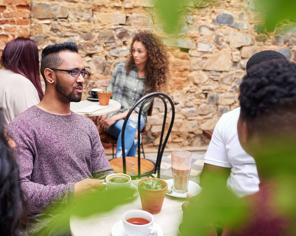 若い男が一緒にトレンディなカフェの中庭にテーブルの周りに座ってコーヒーを飲みながら友人のグループと話して - 写真・画像