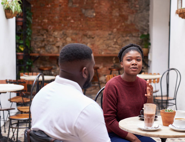 Young African woman talking with a male friend and drinking coffee while sitting together at a table in the courtyard of a trendy cafe - Foto, imagen