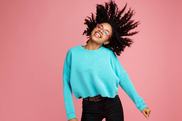 Excited african american young woman with bright smile dressed in casual clothes, glasses and headphones dance over pink background. - Foto, imagen