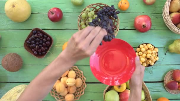 緑の生態学的背景果物。平面図です。緑の木製エコ背景に様々 な果物があります。男性の手は、果物を取るし、赤い板の上に置きます。ここでは、: ブドウ、桃、梅、キウイ、梨、ココナッツ、バナナ、メロン、プラム - 映像、動画