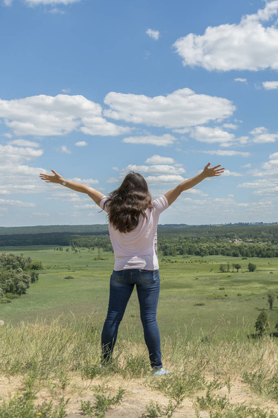 Young hipster girl is travelling in summer holidays. Woman in modern dress is standing on the hill and looking at the field. Blue sky and white clouds. Freedom and lifestyle concept. Beautiful nature. - Foto, afbeelding