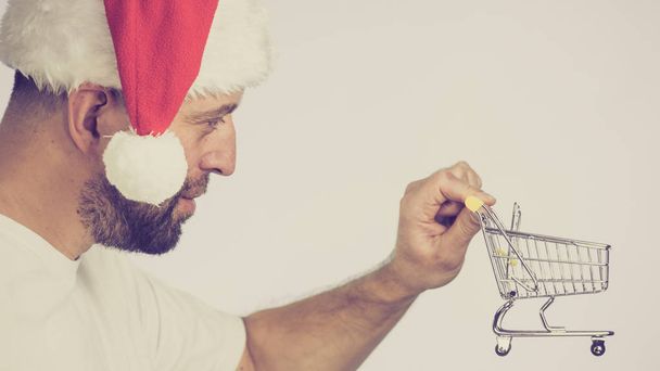 Man wearing santa claus hat holding empty shopping basket cart, toned image. Xmas, seasonal sales, boxing day concept. - Photo, Image