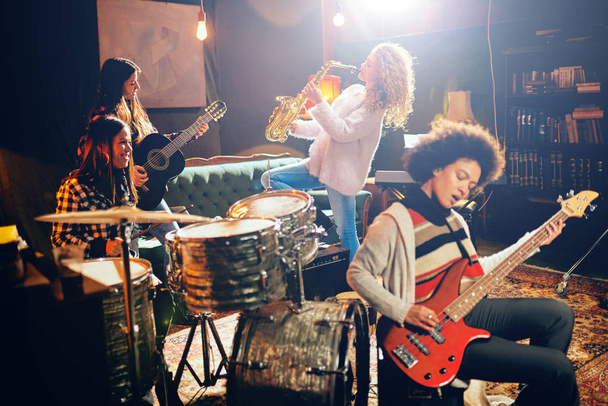 Girls playing jazz music. In foreground one woman playing bass guitar and in background other three playing acoustic guitar, saxophone and drums. Home studio interior. - Photo, Image