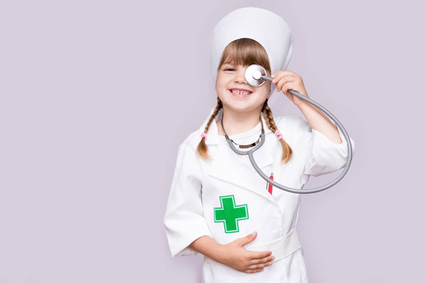 усміхнена дівчинка в медичній формі тримає стетоскоп і дивиться на камеру ізольовано на білому
 - Фото, зображення