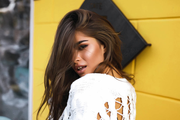 Sexy portrait of young lady with long dark hair falling down on the face. Beautiful makeup and big lips. Yellow background of the cafe. Wearing fashion clothes. Shooting outdoor for the magazine. - Φωτογραφία, εικόνα