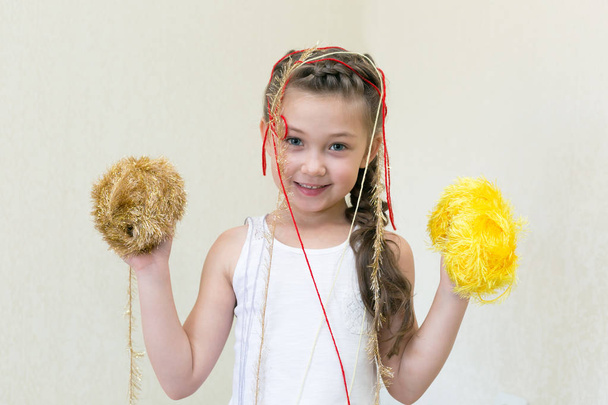 A girl with balls of thread for knitting. The little girl holds knitting yarns in her hands, and the yarn hangs on her head. - Фото, изображение