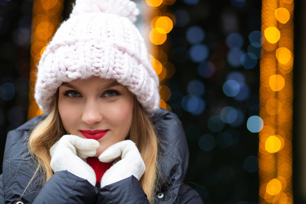 Classy blonde woman with red lipstick wearing knitted cap posing at the street over a garlands background with bokeh - Zdjęcie, obraz