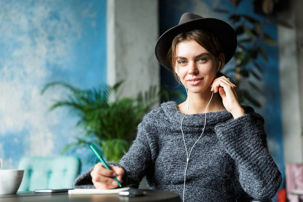 Beautiful smiling young woman dressed in sweater and hat sitting in chair at the cafe table, listening to music with earphones, stylish interior, taking notes - 写真・画像