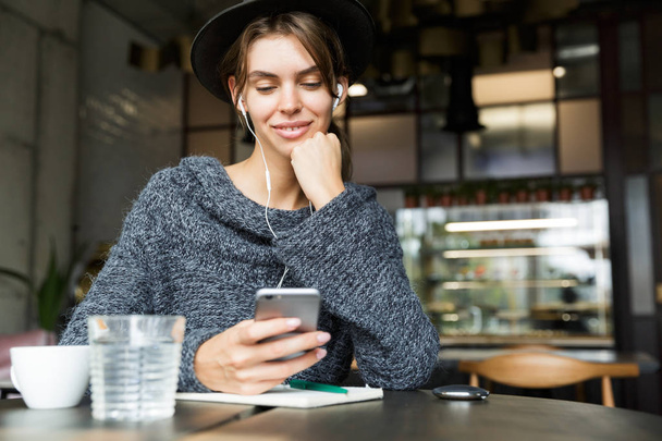 Beautiful lovely young woman dressed in sweater and hat sitting in chair at the cafe table, listening to music with earphones, using mobile phone, stylish interior - Photo, Image
