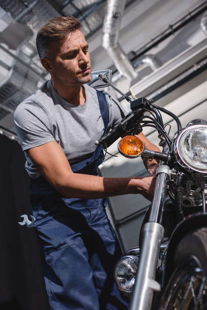 handsome adult mechanic in overalls fixing motorcycle in garage - Фото, изображение
