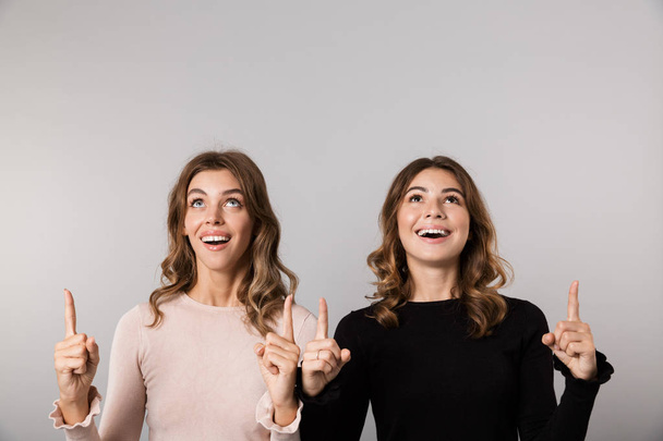 Image of two lovely girls smiling and pointing fingers upward together isolated over gray background - Valokuva, kuva