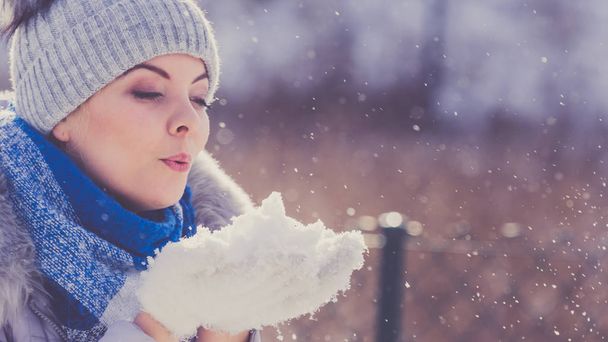 Pretty young woman blowing snow playing with it. Female having grey beanie warm hat with pompons and blue scarf. - Foto, Bild
