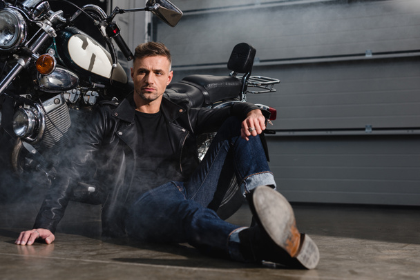 όμορφος άντρας κάθεται στο έδαφος με μοτοσικλέτα στο γκαράζ - Φωτογραφία, εικόνα