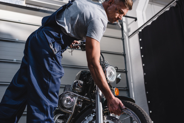 smoking adult mechanic in overalls checking motorcycle tire in garage - Fotografie, Obrázek