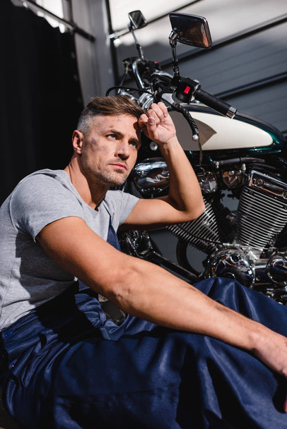mechanic with hand on forehead next to motorbike in garage - Fotoğraf, Görsel