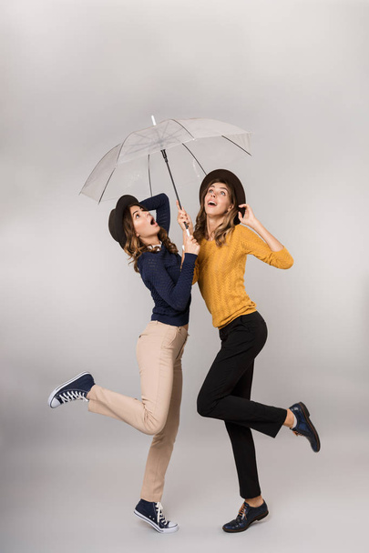 Full length image of two positive girls smiling and walking together under umbrella isolated over gray background - Foto, afbeelding