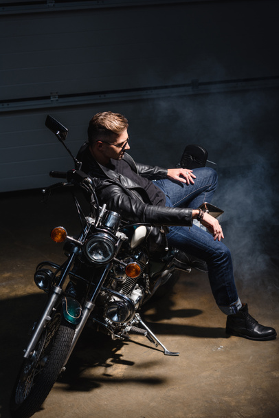 side view of handsome guy in sunglasses sitting on motorcycle in garage - Foto, Imagen