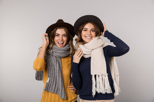 Image of two beautiful women wearing hats and scarfs smiling at camera isolated over gray background - Fotoğraf, Görsel