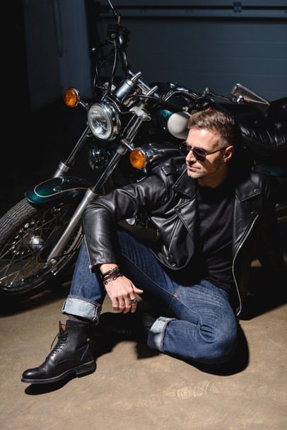 handsome biker in sunglasses sitting by motorcycle in garage - Фото, изображение