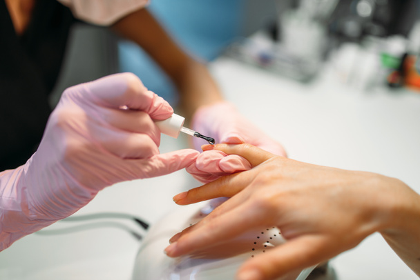 Beautician in pink gloves applying nail varnish to female client, manicure in beauty salon. Manicurist doing hands care cosmetic procedure - Foto, Imagem
