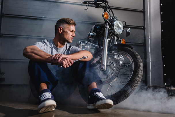 mecánico guapo mirando hacia otro lado, sosteniendo la llave inglesa y sentado cerca de la motocicleta en el garaje
  - Foto, Imagen