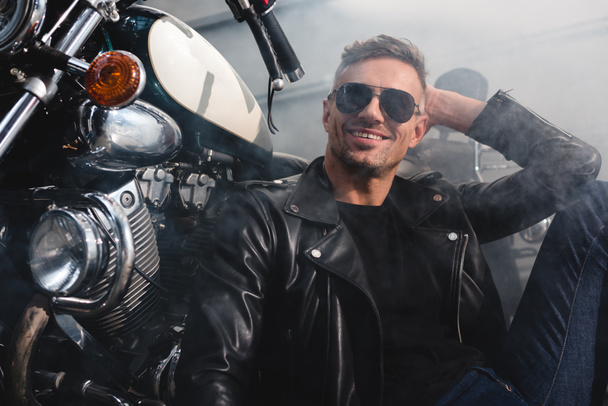 handsome man in sunglasses smiling sitting by motorcycle in garage - Zdjęcie, obraz