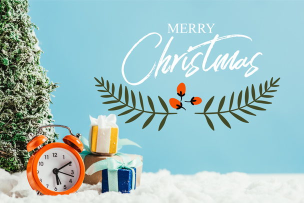 close-up shot of christmas gifts with alarm clock and miniature christmas tree standing on snow on blue background with "merry christmas" lettering with leaves illustration - Foto, imagen