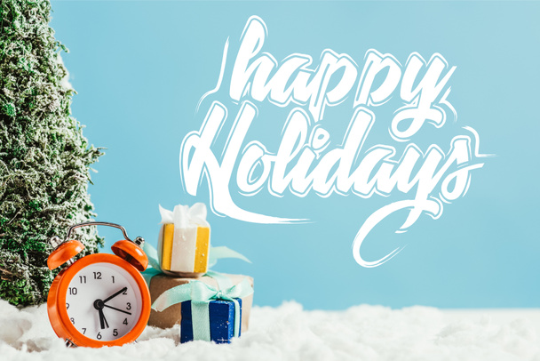Detailní záběr vánočních dárků s Budík a miniaturní vánoční strom stojící na sněhu na modrém podkladu s "happy holidays" nápisy - Fotografie, Obrázek
