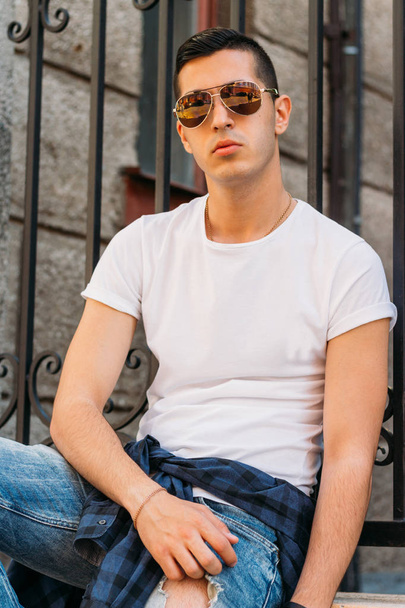 Attractive, sexy guy sits leaning on a curb on a ledge in the wall on the street in sunglasses. self-confidence, flatulence and arrogance.model, posing, portrait of a young man - Foto, Imagen