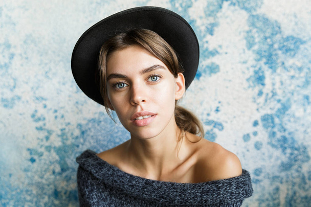 Close up portrait of a young woman dressed in sweater and hat over blue wall background, looking at camera - Photo, Image